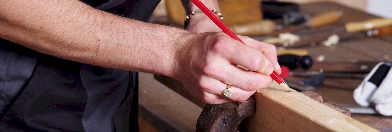 Das Bild zeigt eine menschliche Hand, die einen Zimmermannsbleistift hält und auf einem Stück Holz eine Markierung anbringt. Im Bildhintergrund ist die Tischplatte eines Werktischs zu sehen auf der, unscharf dargestellt, verschiedene Holzwerkzeuge liegen