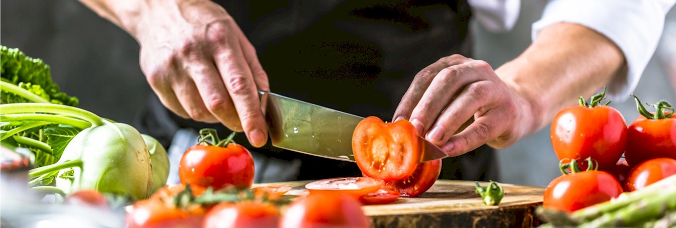 Das Bild zeigt zwei menschliche Hände. Die eine Hand hält ein großes Küchenmesser und schneidet eine Tomate, welche von der anderen Hand gehalten wird. Vor und neben den Händen liegen weitere Tomaten, ein Kolrabi und grüner Spargel.