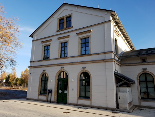 Das Bild zeigt das Gebäude des Vereins am Standort Oelsnitz. Es ist ein ehemaliges Bahnhofsgebäude. Das Haus ist weiß verputzt, zweistöckig und hat Sprossenfenster. Die Fenster im Erdgeschoß und die Tür sind als Rundbogen gebaut.